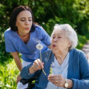 elderly woman blowing out a dandelion clock with help of a carer